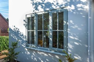 White uPVC Casement windows on a sunlit wall with dappled shadows from an overhead tree, offering a glimpse into the warm interior and garden beyond.