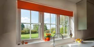 The image showcases a bright kitchen with a large uPVC Casement window that frames a serene garden view. The window, with its clean white frames, is divided into multiple panes, allowing ample natural light to flood the room. A bold orange roller blind adds a splash of vibrant color at the top, complementing the outdoor greenery. On the windowsill, there's a cheerful display of orange flowers in a matching pot, and a bowl of fresh lemons adds to the zestful atmosphere. The sink faucet reflects the sunlight, and overall, the scene is one of warmth and domestic charm.