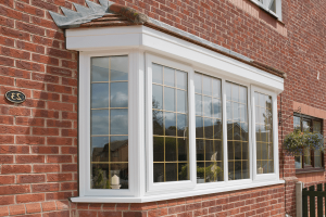 A white uPVC bay window with grid designs installed in a red brick house, reflecting a clear sky and the surrounding greenery, with a hanging flower basket adding a touch of charm to the quaint exterior.