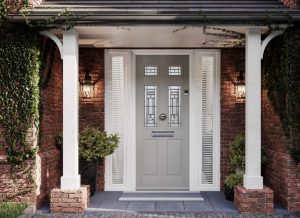 The image shows a front porch of a brick house with a white wooden canopy supported by two white columns. The grey front door has decorative glass panels and a mail slot, flanked by narrow windows on either side. A wall-mounted lantern light is attached to the brick wall beside the door. The porch area is adorned with green climbing plants and potted shrubs, creating an inviting entrance.